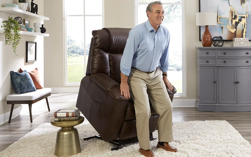A smiling man uses the lift function of a brown leather medical power lift chair to stand up safely and easily in his well-lit, comfortable living room.