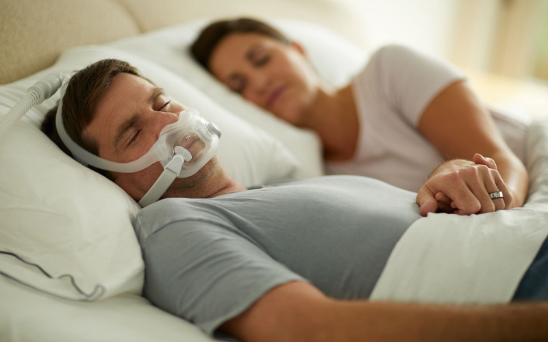 Man sleeping with CPAP mask in bed holding woman's hand