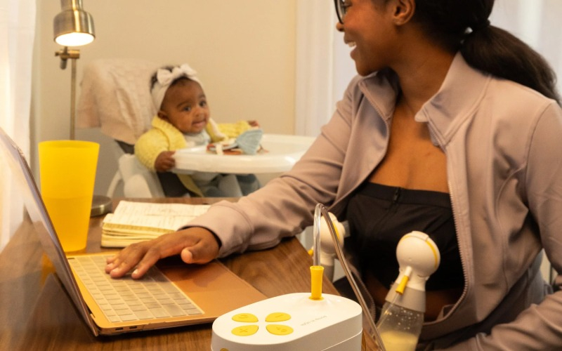 A smiling Black mother sitting at a wooden desk, working on a laptop while using the Medela Pump In Style Pro breast pump hands-free. In the background, a happy baby in a yellow sweater sits in a high chair, illustrating the seamless balance of profession