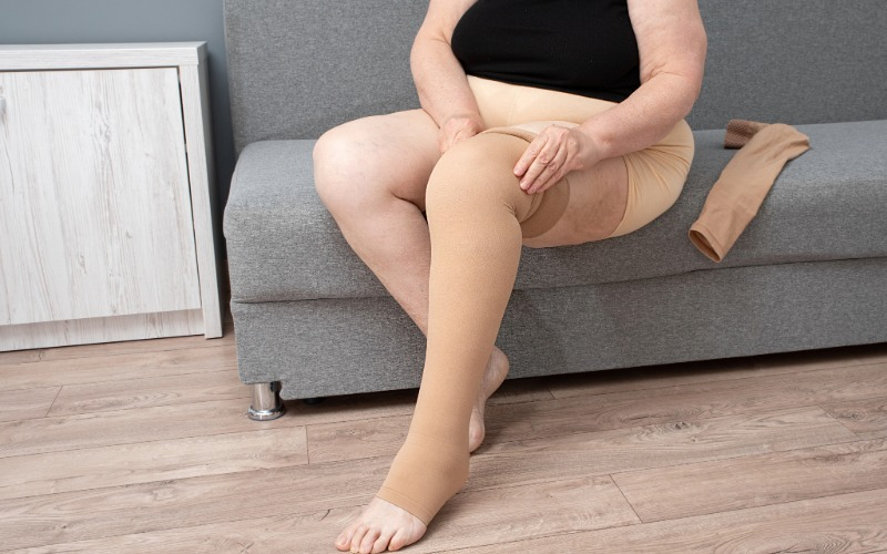 A person sits on a gray couch and pulls a beige, thigh-high compression stocking onto their leg to manage symptoms of Chronic Venous Insufficiency.
