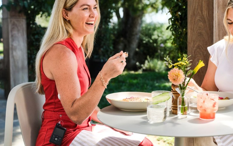 A smiling woman enjoying brunch at a dining table, with a diabetes wearable device visible on her arm.