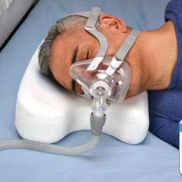 Side sleeper wearing a full face CPAP mask resting on a CoolPAP pillow with cutouts to accommodate the mask tubing, alongside an inset graphic highlighting the blue cooling gel layer for temperature control.