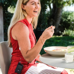 A smiling woman enjoying brunch at a dining table, with a diabetes wearable device visible on her arm.