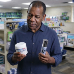 A middle-aged Black man stands in a Binson's retail store holding two skin repair products, carefully comparing the labels on a jar of Deep-Healing Foot Cream and a bottle of K5 Neuropathy Relief & Skin Repair Cream.