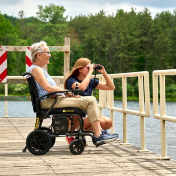 An elderly woman smiling in a black Golden Cricket LT power wheelchair on a wooden dock alongside a girl looking through binoculars over the water.
