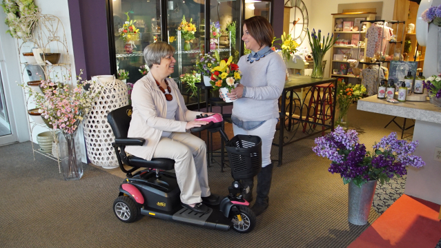 Woman smiling on a pink Golden Technologies Buzzaround EX 3-wheel mobility scooter while receiving flowers from a florist.