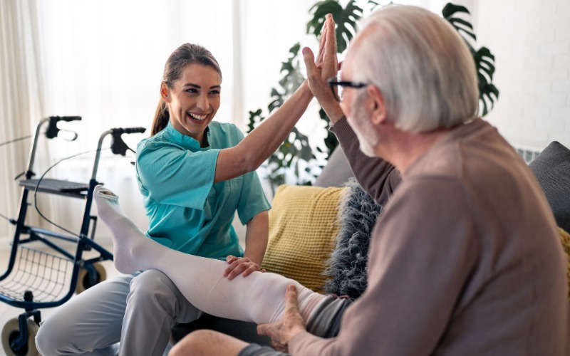 A smiling female healthcare professional and an older male patient give each other a high-five as she helps him put on a white compression stocking in his home.