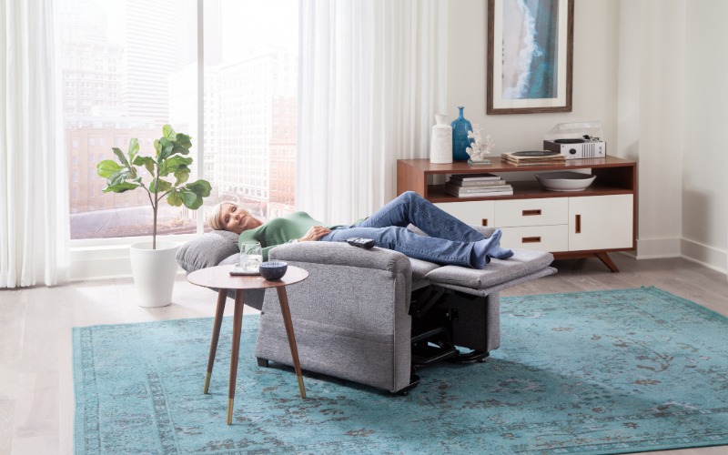 A woman relaxes comfortably in a fully reclined grey fabric medical lift chair with her feet elevated, demonstrating the chair's therapeutic support in a bright, modern living room.